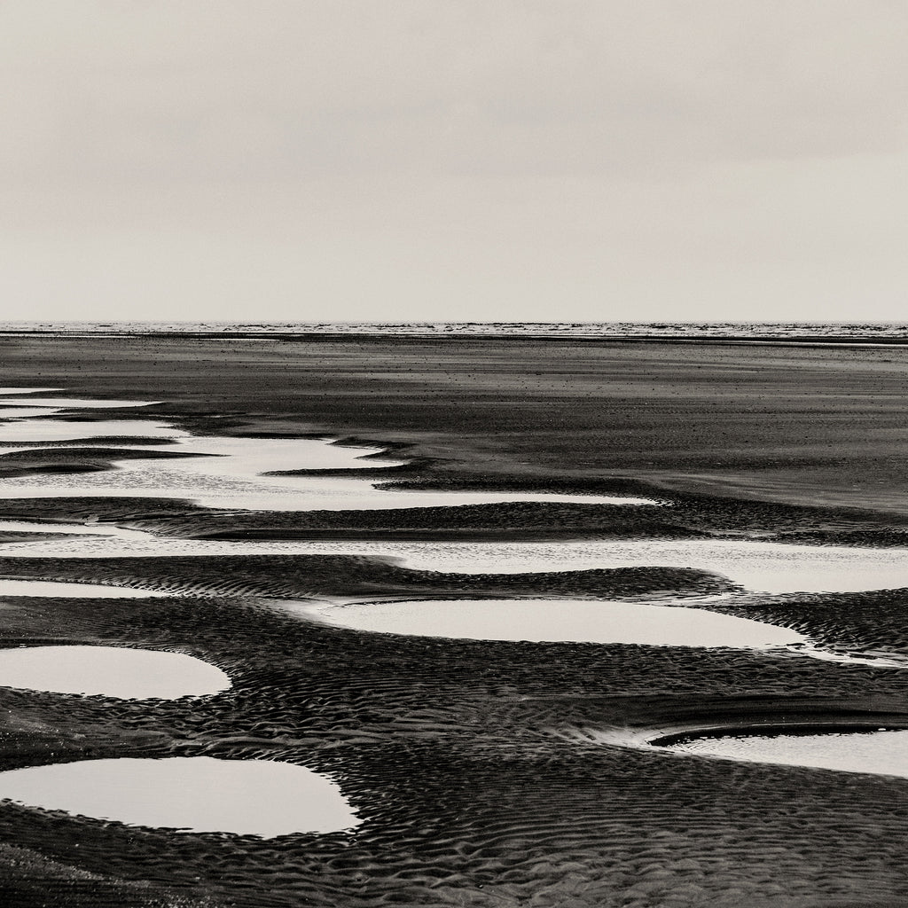 Black and white landscape photograph of a tidal flat or beach with shallow pools of water reflecting the sky, shown as a fine art print.