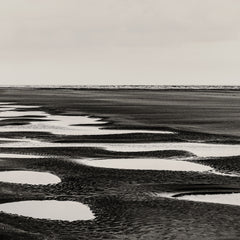 Black and white landscape photograph of a tidal flat or beach with shallow pools of water reflecting the sky, shown as a fine art print.